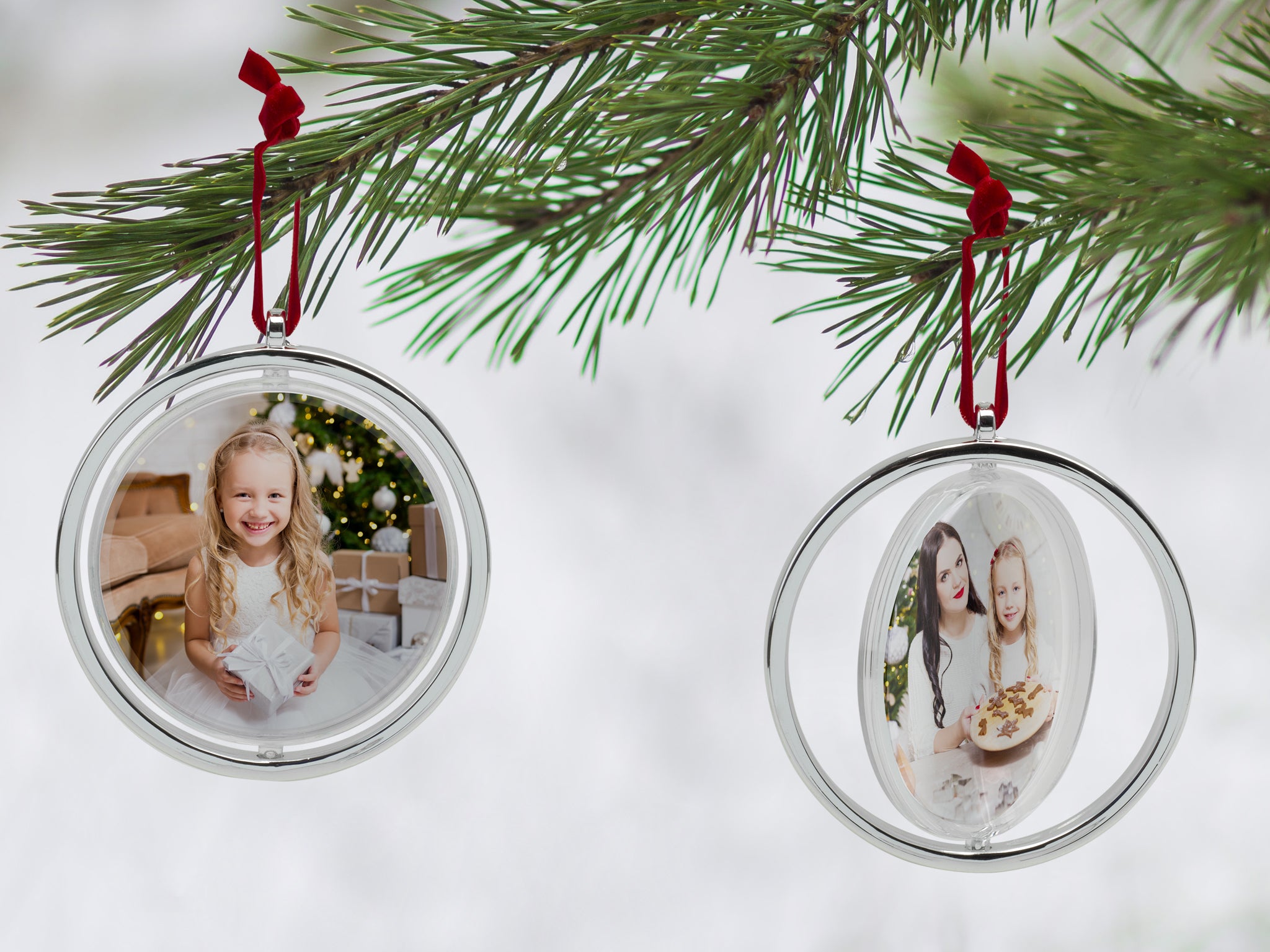 Two round, silver photo ornaments with a red velvet string at the top hanging on a Christmas tree.