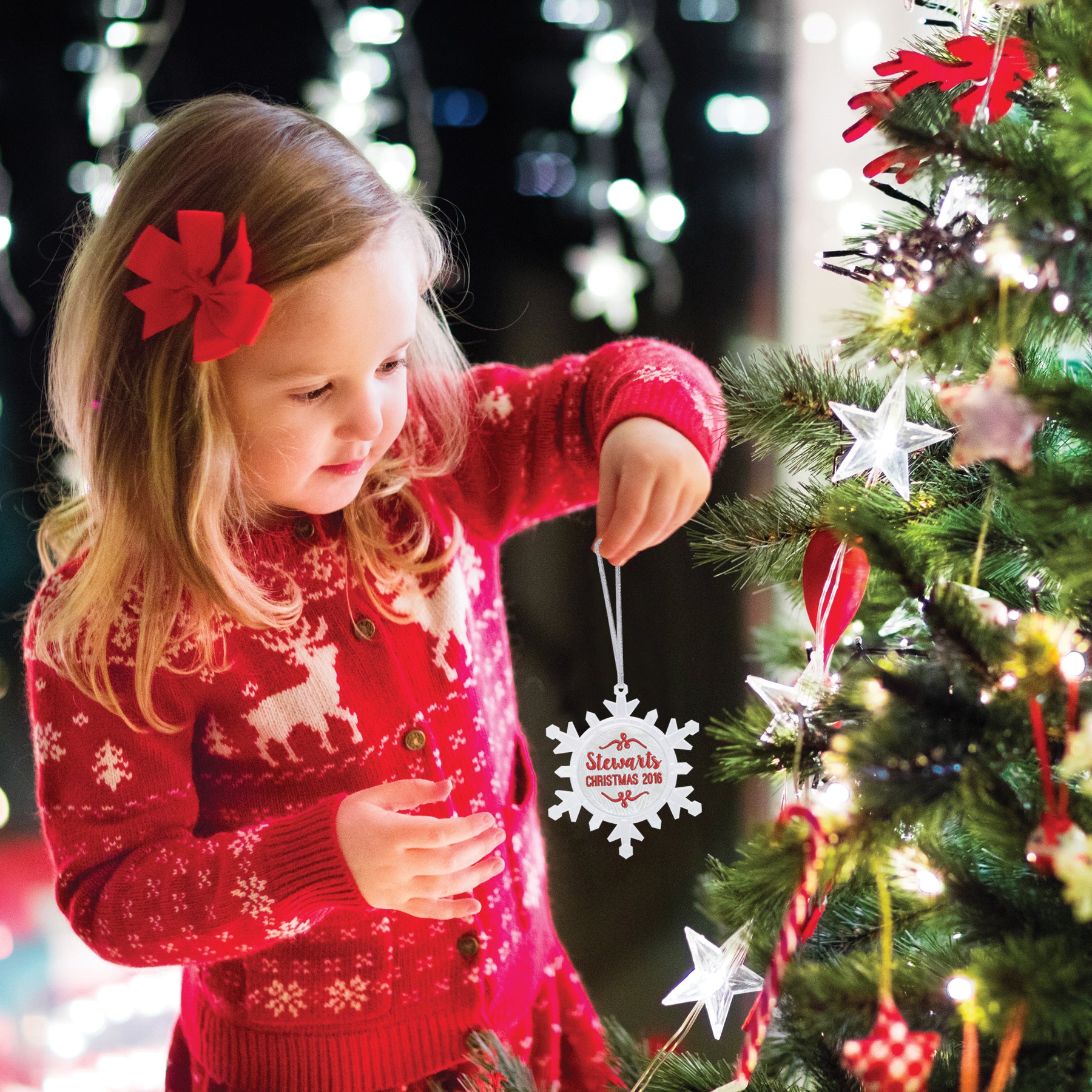 A clear plastic snowflake-shaped photo ornament with a beveled edge. The ornament is designed to hold a photo or embroidery design, and has red/green/blue LED lights and is shown being held by a young child about to hang it on a Christmas tree in a festive lifestyle background.