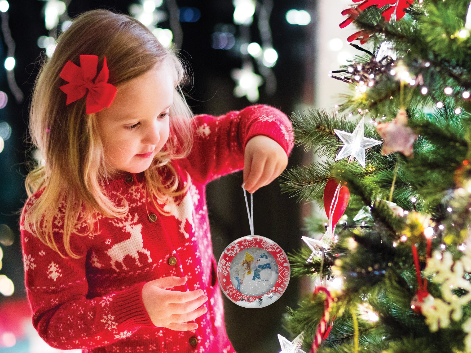 A round snow globe photo ornament with a red border and white snowflakes being help by a young girl about to put it on a Christmas tree.