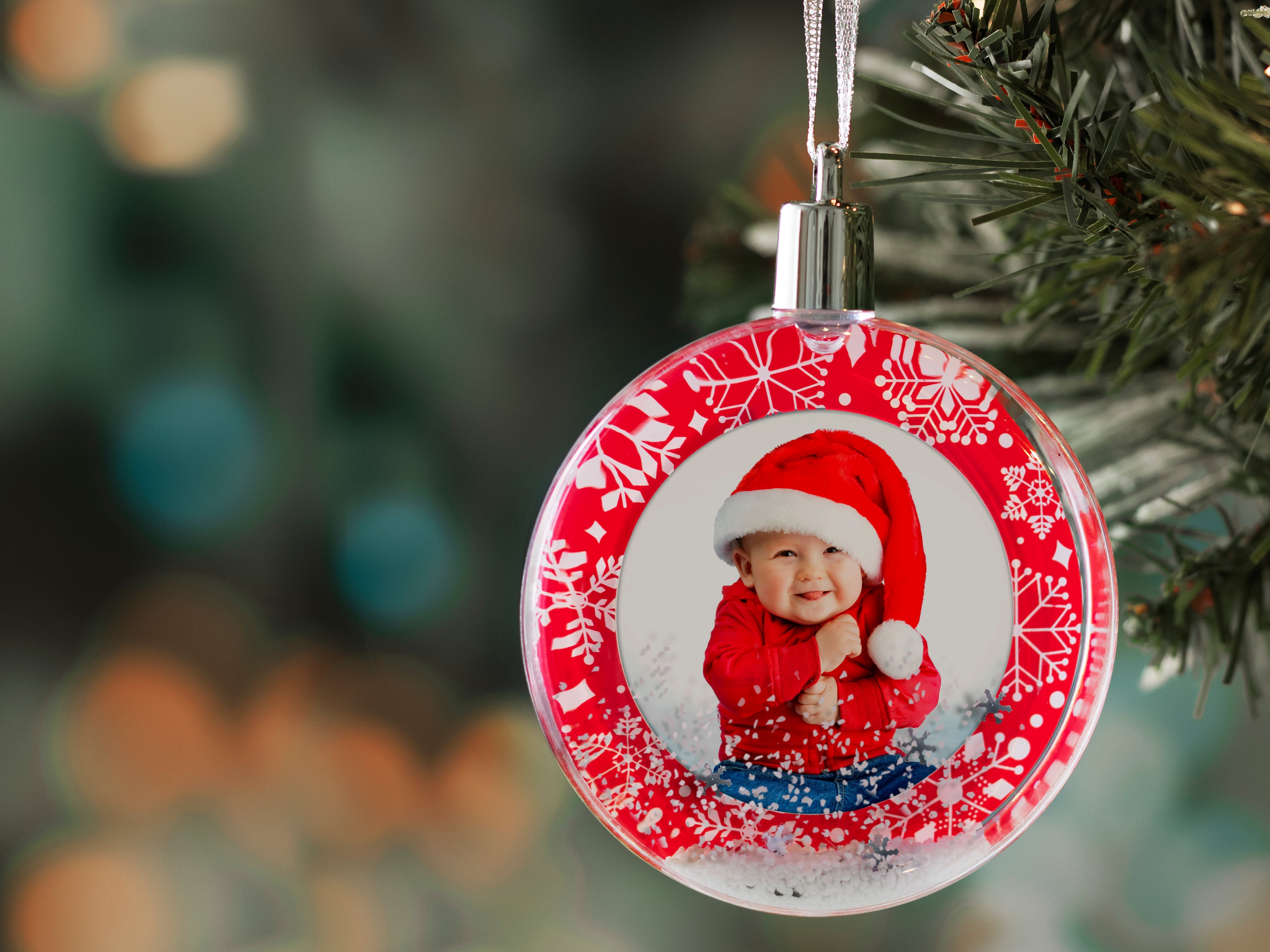 A round snow globe photo ornament with a red border and white snowflakes with a silver ribbon and cap hanging on a Christmas tree on a festive lifestyle background.