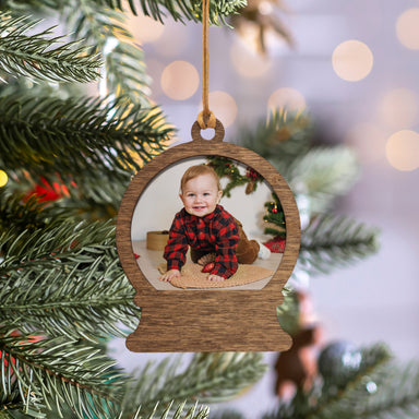 A wooden snow globe-shaped photo ornament with a picture of a baby inside, hanging by a raffia string on a decorated Christmas tree.