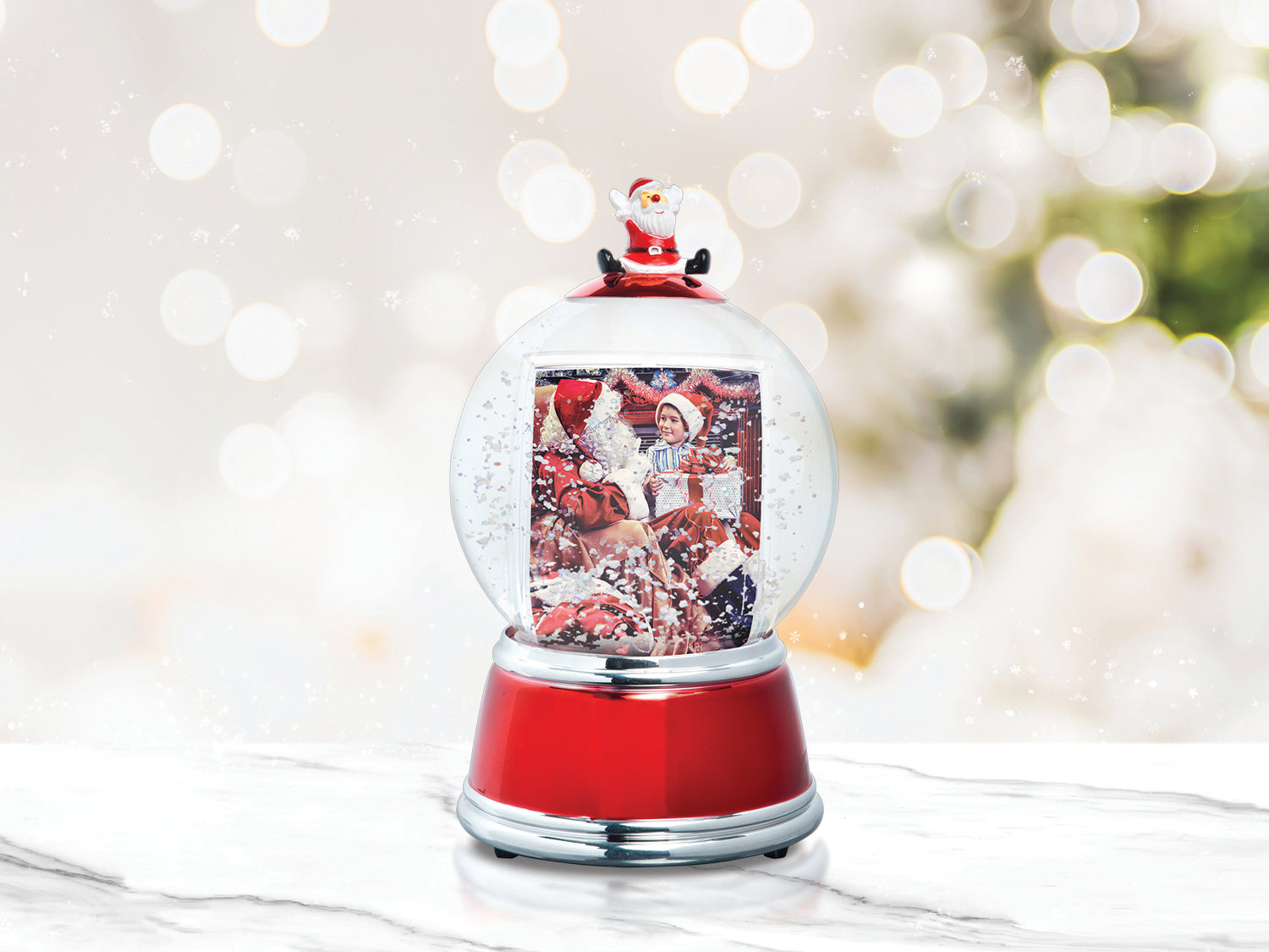 A holiday themed photo snow globe with LED lights. The globe is clear with a Santa figurine on the top, and the base is red with silver trim displayed on a festive lifestyle background.