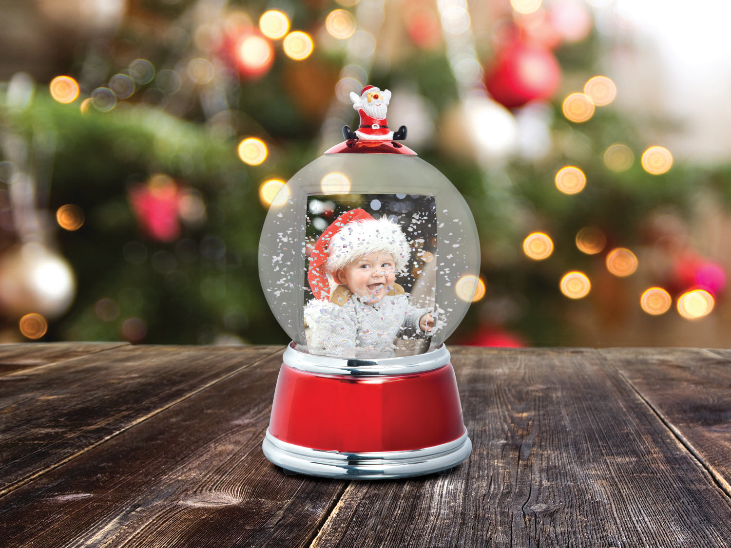 A holiday themed photo snow globe with LED lights. The globe is clear with a Santa figurine on the top, and the base is red with silver trim displayed on a festive lifestyle background.