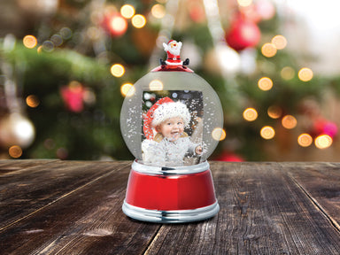 A holiday themed photo snow globe with LED lights. The globe is clear with a Santa figurine on the top, and the base is red with silver trim displayed on a festive lifestyle background.