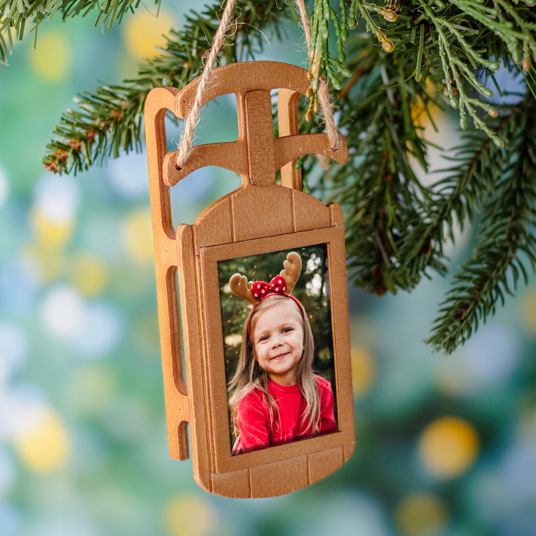 A wooden sled-shaped photo ornament seen hanging on a Christmas tree by a loop of twine.