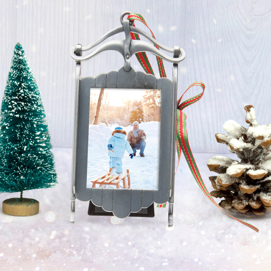A pewter sled-shaped photo ornament and picture frame adorned with a red, green, and gold ribbon displayed on table in a festive lifestyle background.