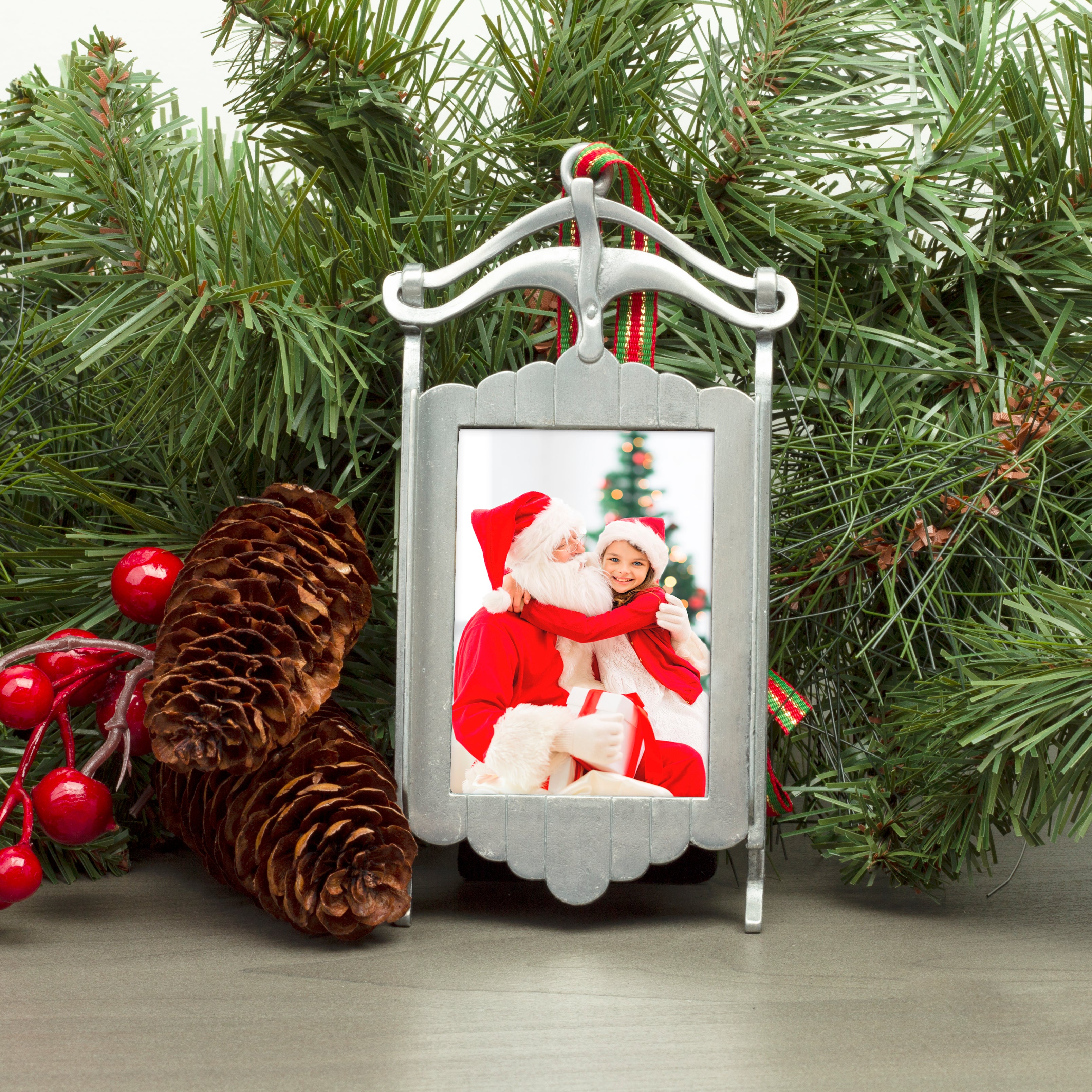 A pewter sled-shaped photo ornament and picture frame adorned with a red, green, and gold ribbon displayed on a table in a festive lifestyle background.