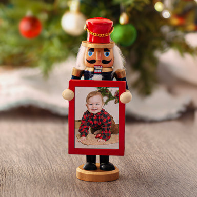 A hand-painted mini photo nutcracker with a gold base, dressed in a navy blue coat with gold accents, red pants, and black boots, holding a photo frame with  Christmas tree background.
