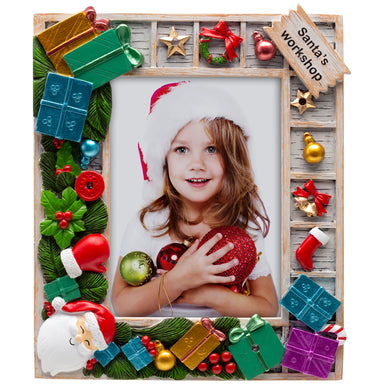 A festive Santa's Workshop resin picture frame with hand-painted details, featuring green garland, mistletoe, wrapped presents, and ornaments. The frame holds a photo of a smiling child wearing a Santa hat, with a light-up border.