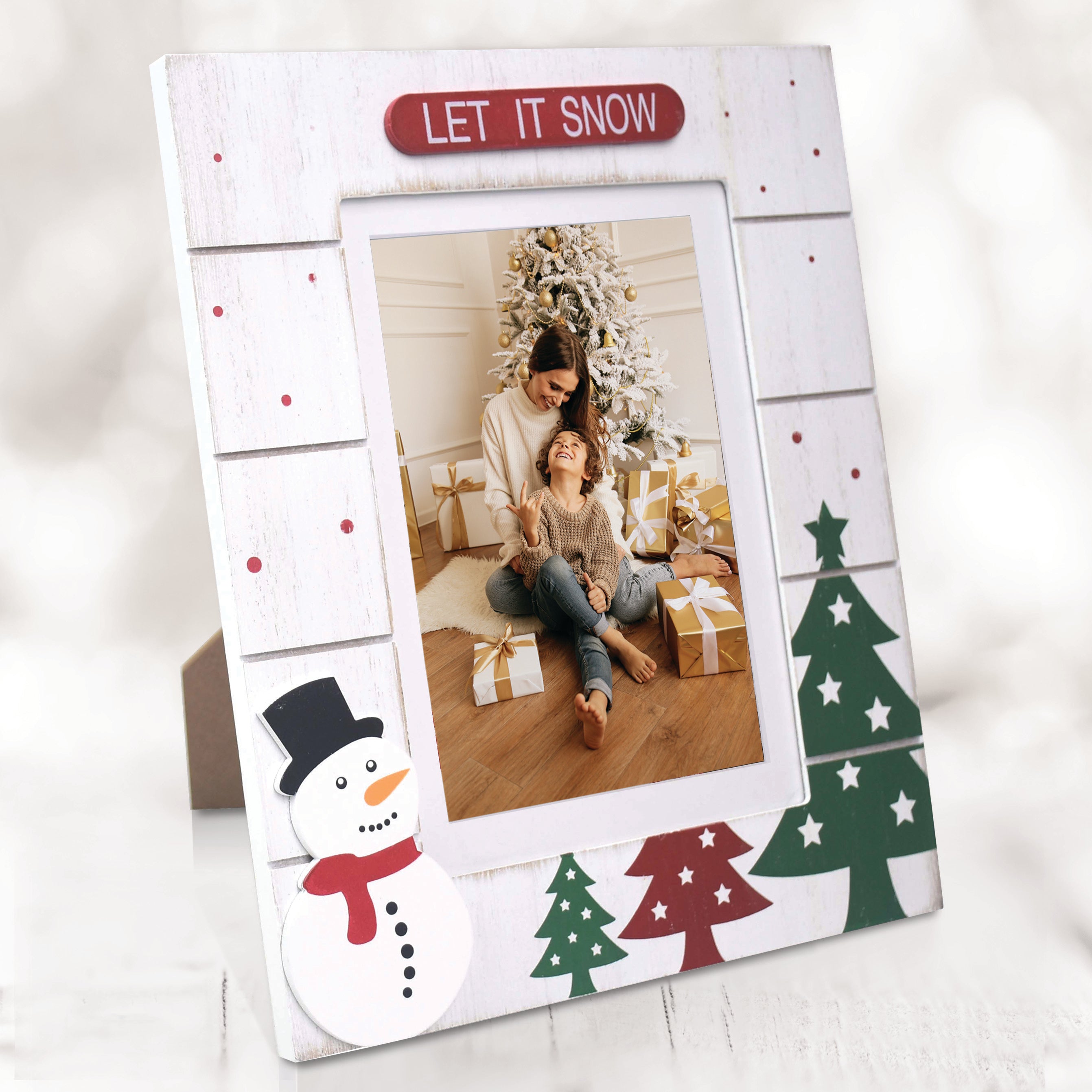 A white, wooden picture frame with a 'LET IT SNOW' sign at the top, and designs featuring Christmas trees, and and a snowman on a festive lifestyle background.