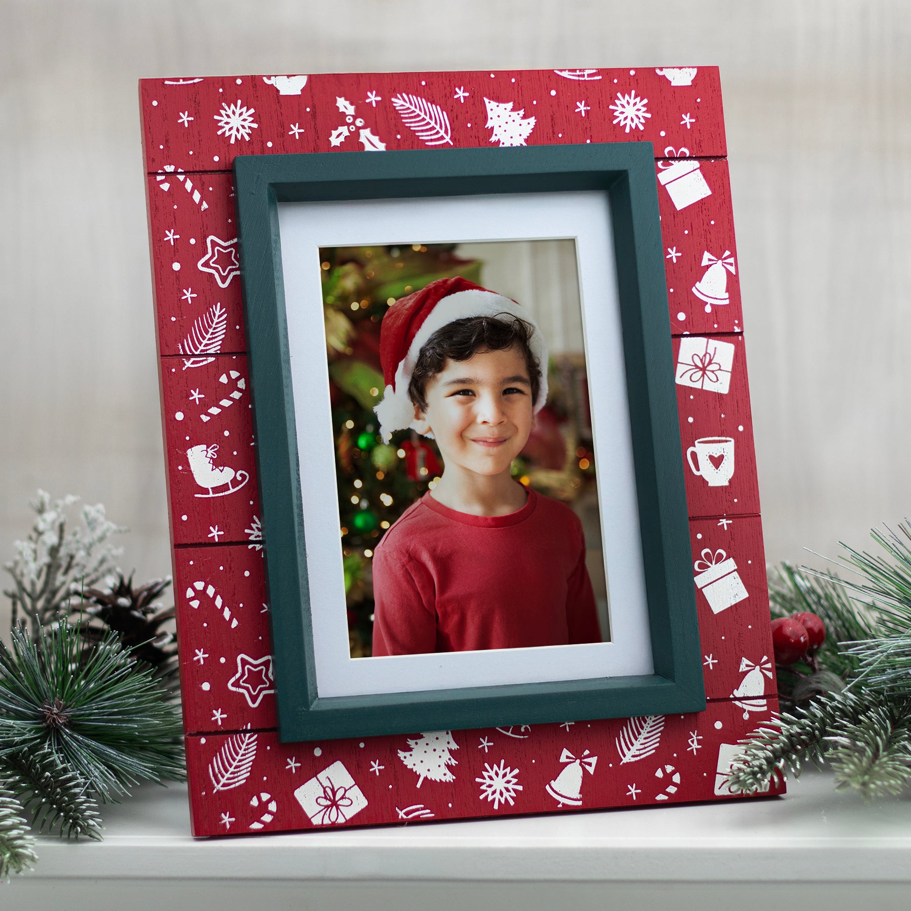 A photo of a child wearing a Santa hat, framed in a red wood picture frame with black trim, displayed against a Christmas-themed backdrop with holiday decorations.