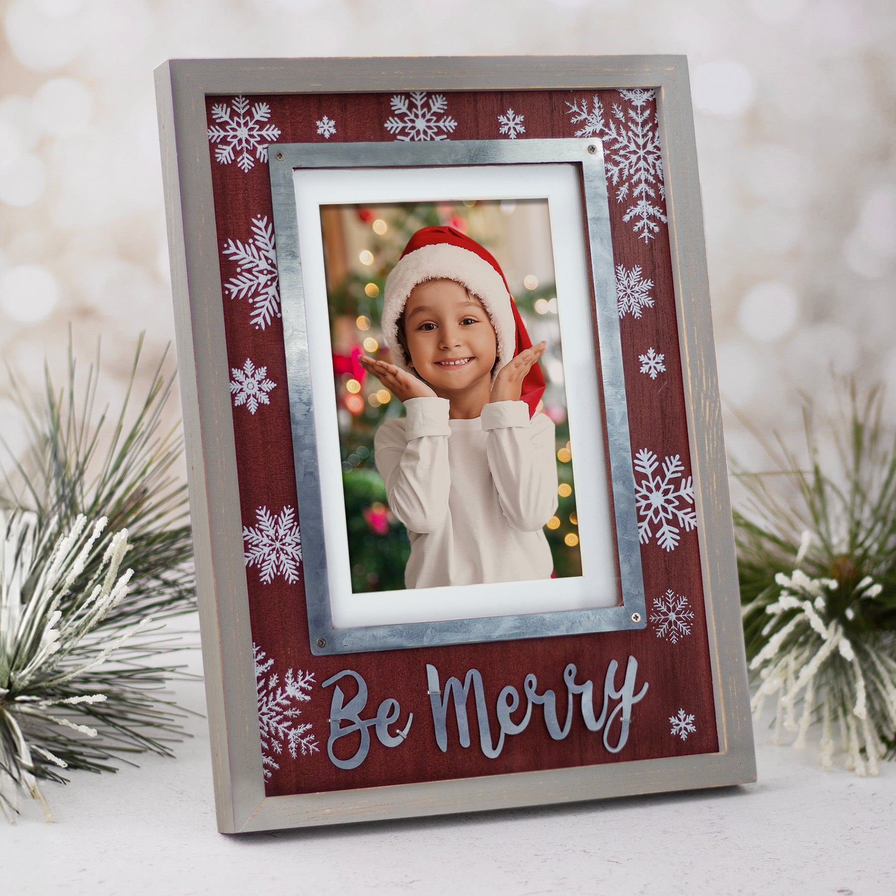 A Christmas-themed wooden picture frame with a gray border and reddish/brown insert. It features white snowflakes in varying sizes in the background and the phrase 'Be Merry' in silver at the bottom shown with a festive back drop.