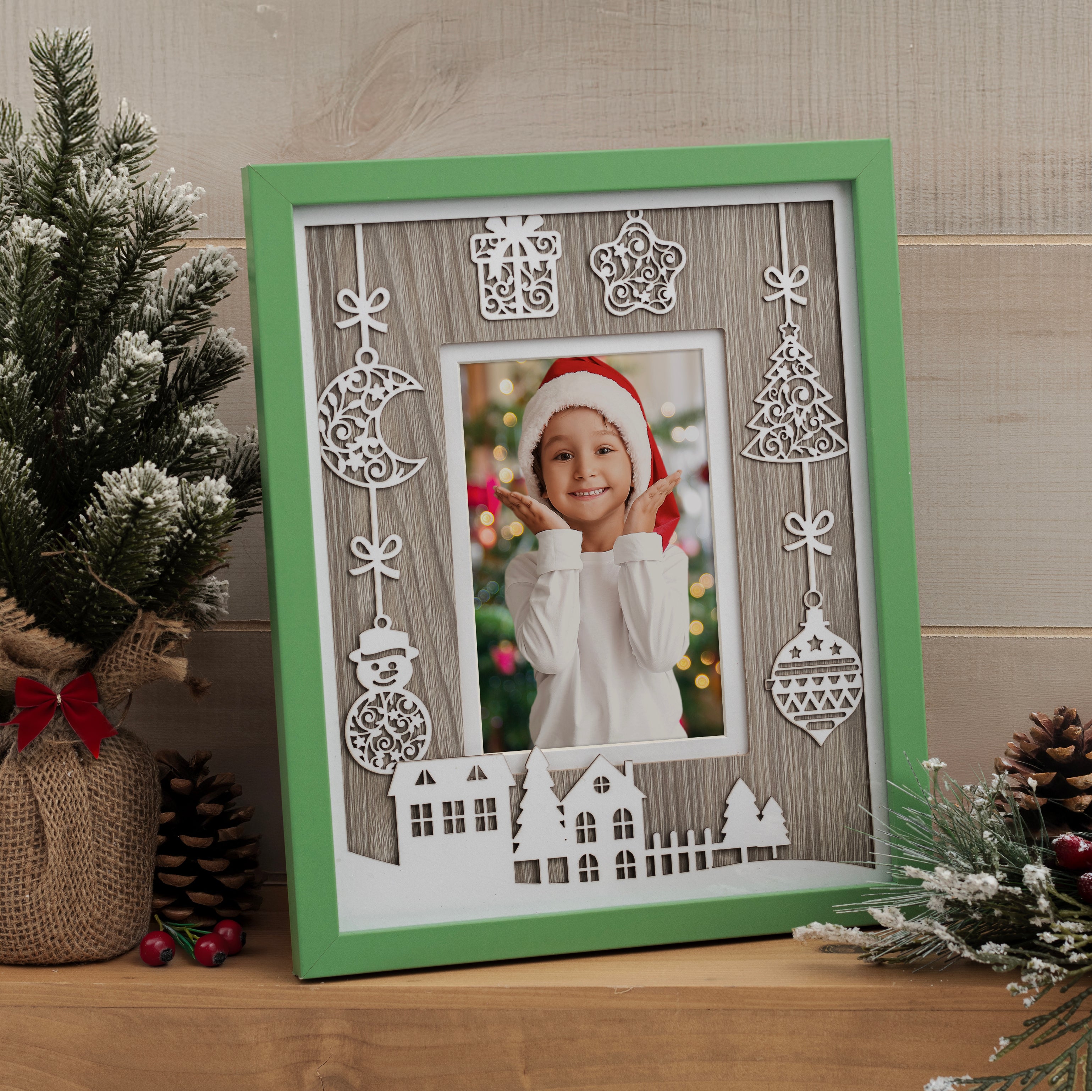 Photo of a green Christmas picture frame with white laser cut designs such as presents, ornaments, and trees shown in a house setting on a mantel or shelf.