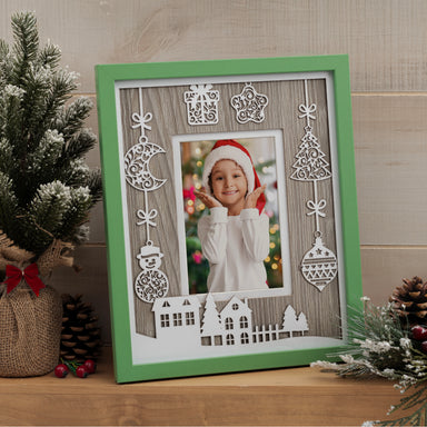 Photo of a green Christmas picture frame with white laser cut designs such as presents, ornaments, and trees shown in a house setting on a mantel or shelf.