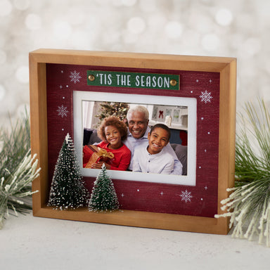 A wooden picture frame with a red background featuring white snowflakes and the text 'Tis the Season'. The frame contains a photo of a family and has two 3D Christmas trees at the bottom displayed with a festive backdrop.