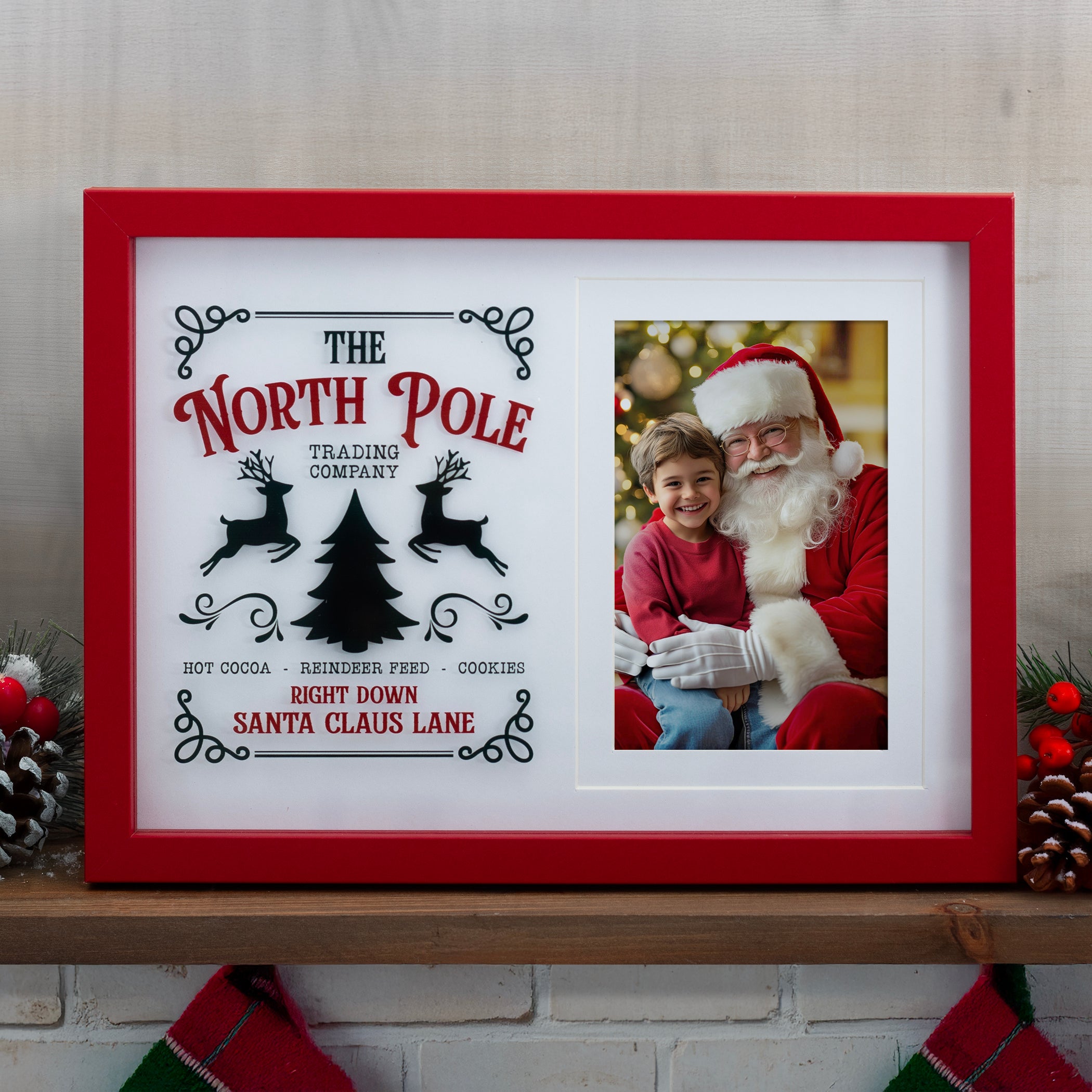 A wooden picture frame with a red border, featuring a photo of a young girl and Santa Claus, and text that reads 'The North Pole Trading Company, Hot Cocoa, Reindeer Food, Cookies, Santa Claus Lane'. Photo is shown standing in a home on a shelf or on a mantel.