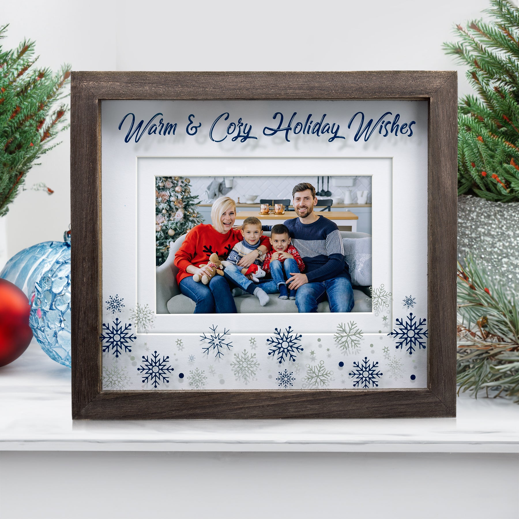A holiday-themed picture frame with a brown border and white inner border, featuring snowflakes, dots, and stars in blue and silver at the bottom sitting on top of a mantel.