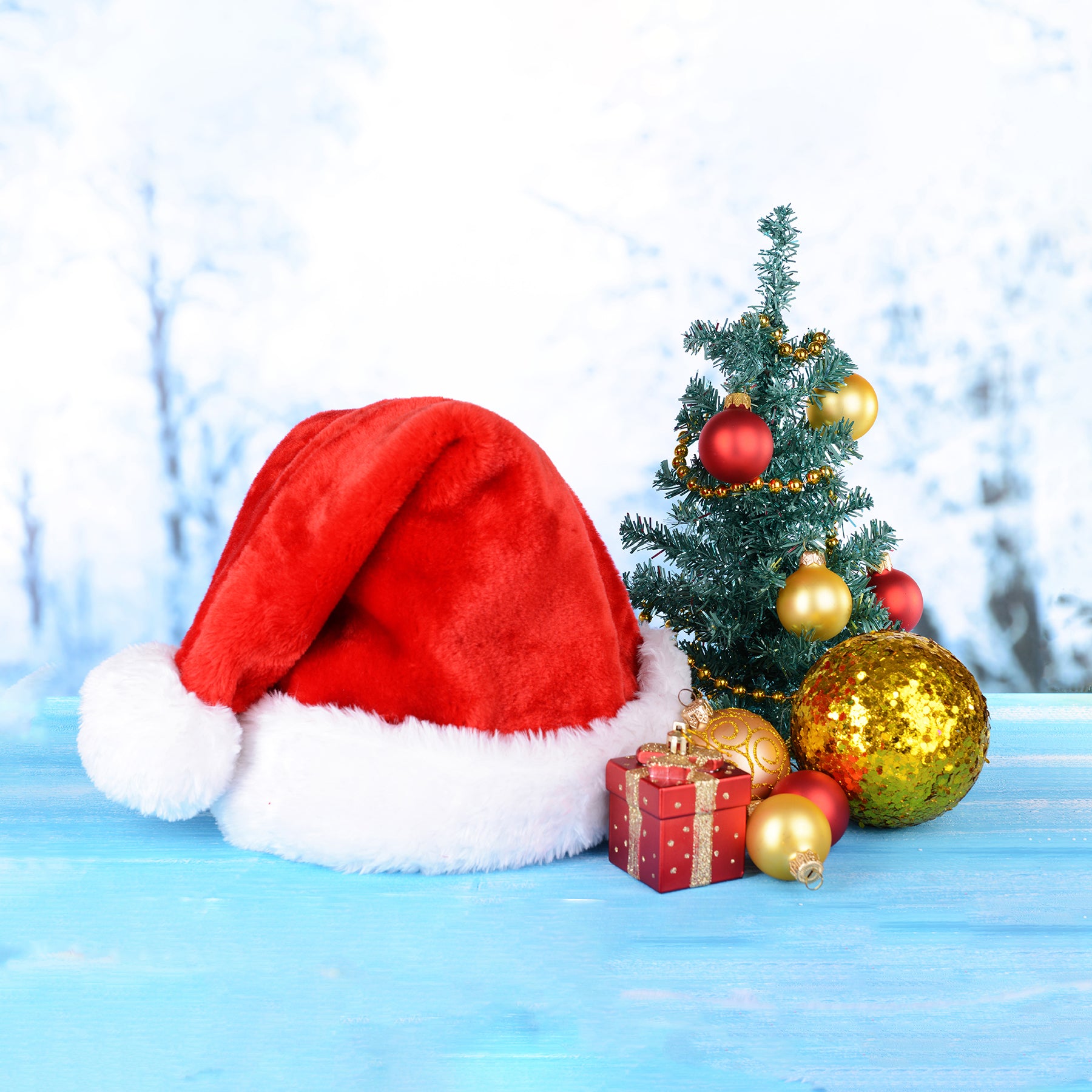 A red Santa hat with white trim next to a mini Christmas tree with ornaments, and a present.
