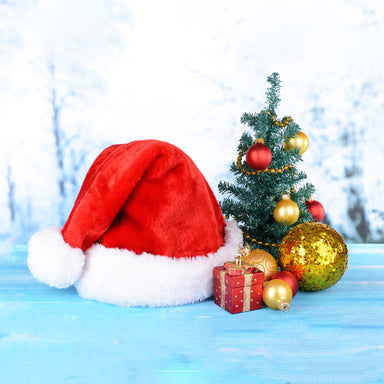 A red Santa hat with white trim next to a mini Christmas tree with ornaments, and a present.