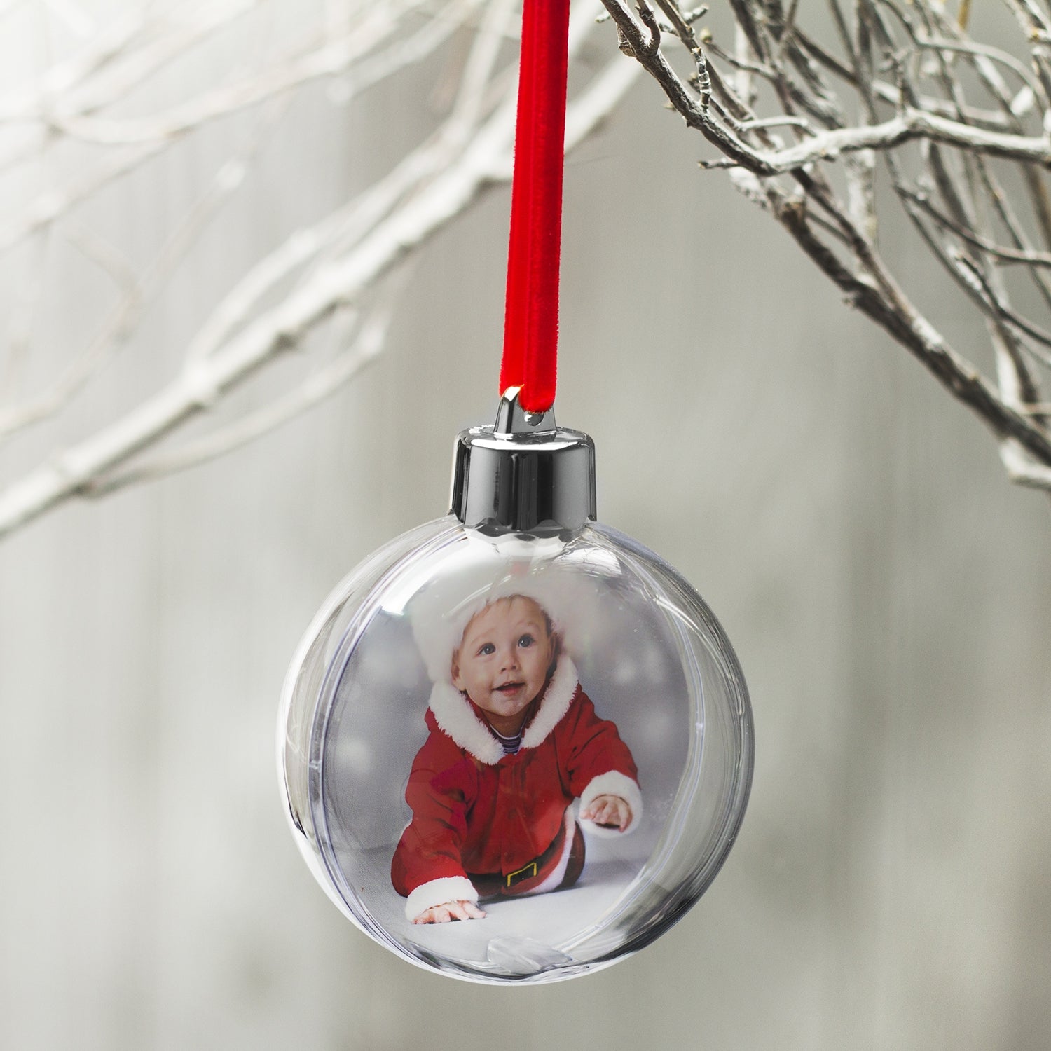 A clear, round Christmas ornament with a photo of a smiling child in Santa attire, and a red velvet hanger ribbon hanging on a tree displayed on a festive lifestyle background.