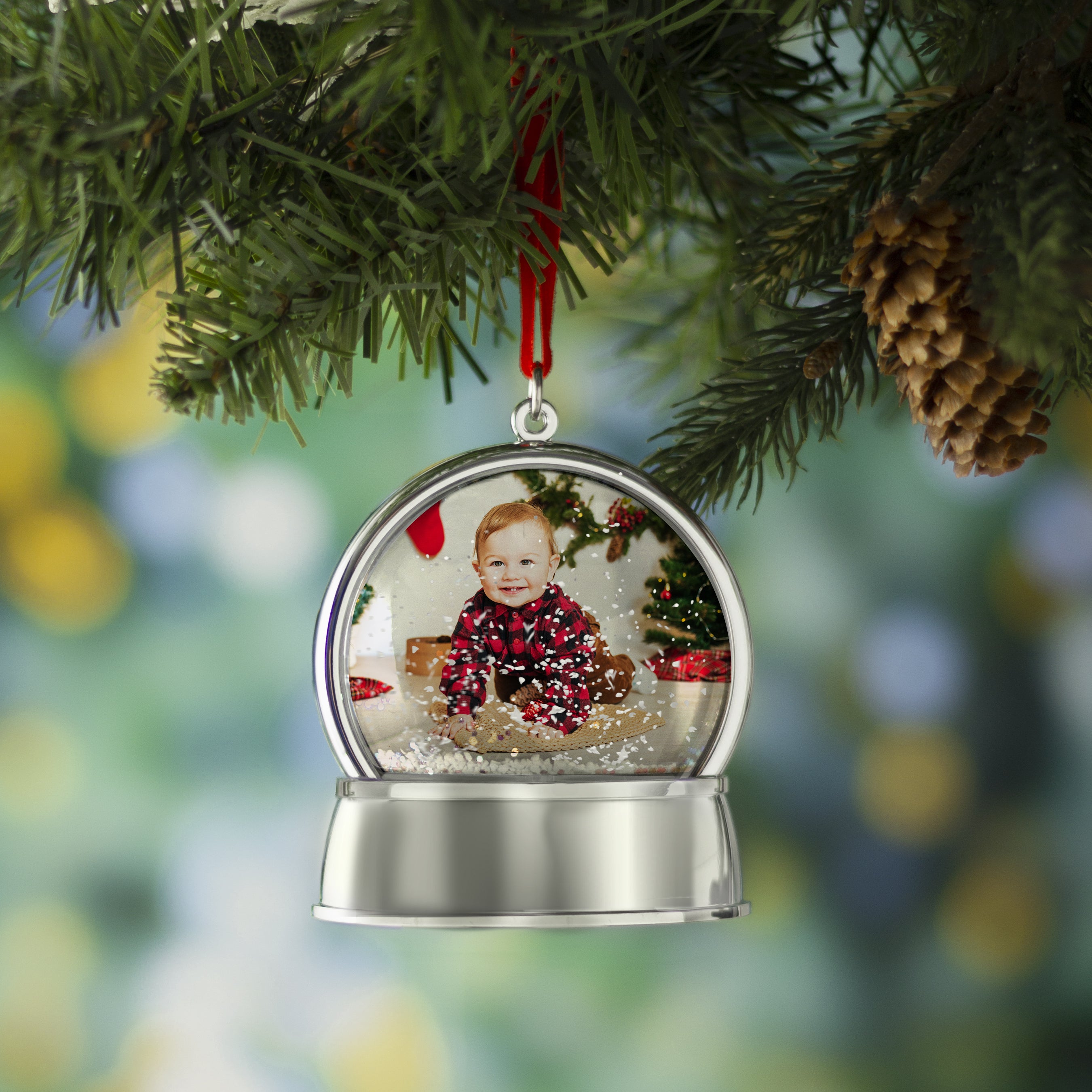 A magnetic snow globe ornament with a silver base and a curved photo inside the globe hanging on a Christmas tree by a red velvet ribbon displayed on a festive lifestyle background.