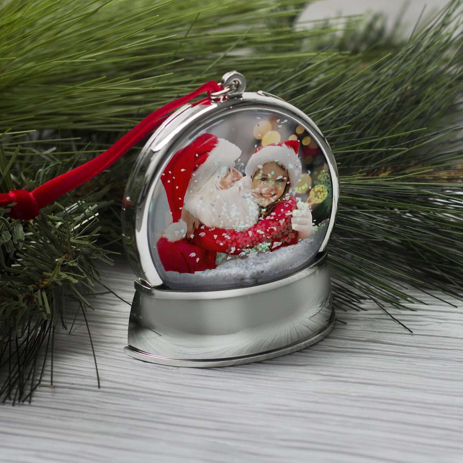 A magnetic snow globe ornament with a silver base and a curved photo of two smiling children wearing Santa hats inside the globe. The ornament has a red velvet string for hanging and features white snow and iridescent glitter being displayed on a table in a festive lifestyle background.