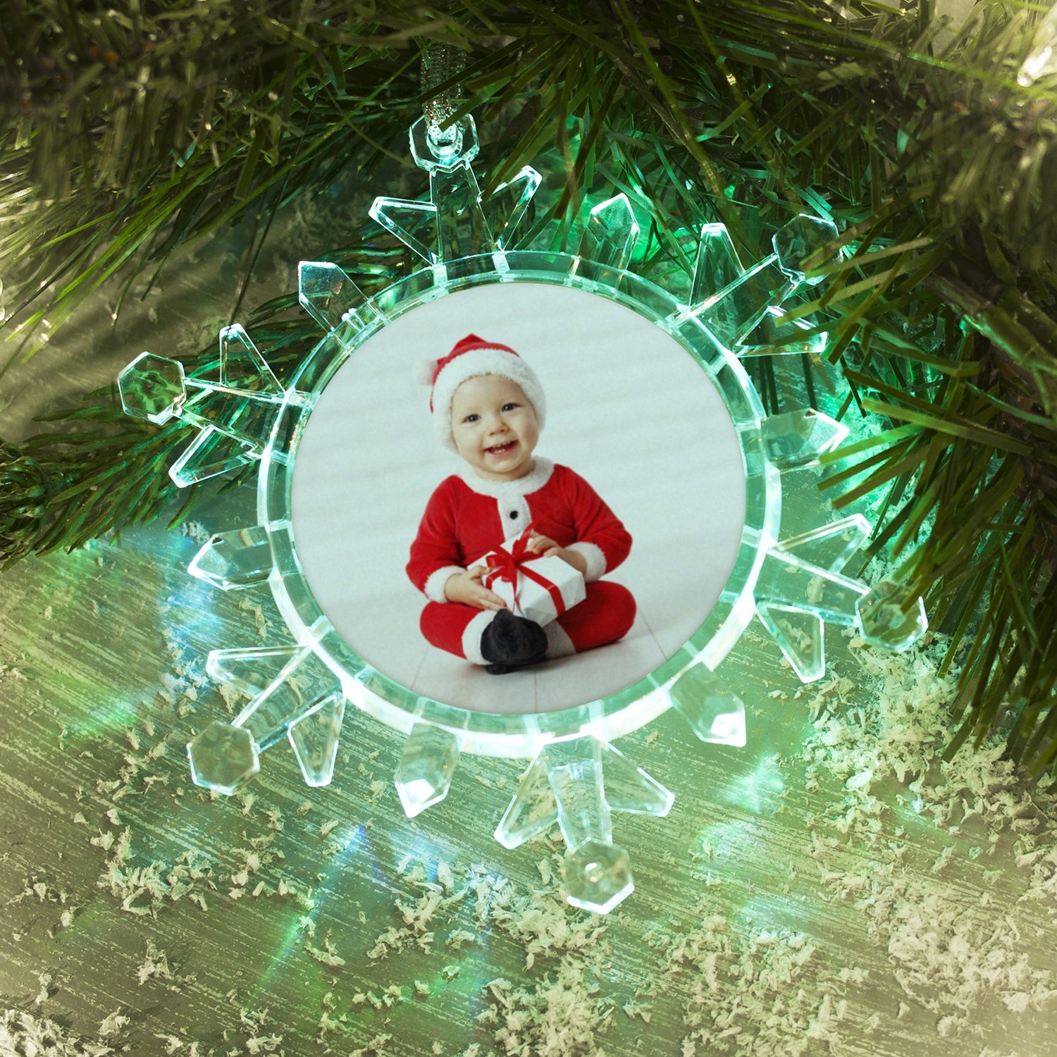 A clear plastic snowflake-shaped photo ornament with a beveled edge, displaying a photo of a smiling child in Santa attire in the center. The ornament is showcasing green LED lights, laying on a table on a festive lifestyle background.