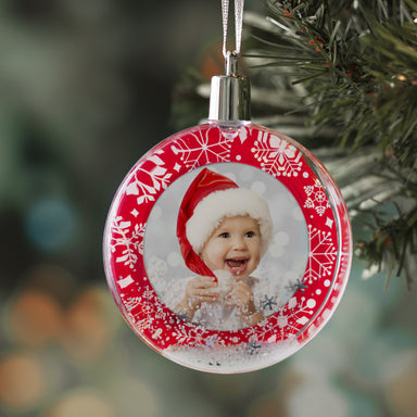 A round snow globe photo ornament with a red border and white snowflakes with a silver ribbon and cap, hanging on a Christmas tree on a festive lifestyle background.