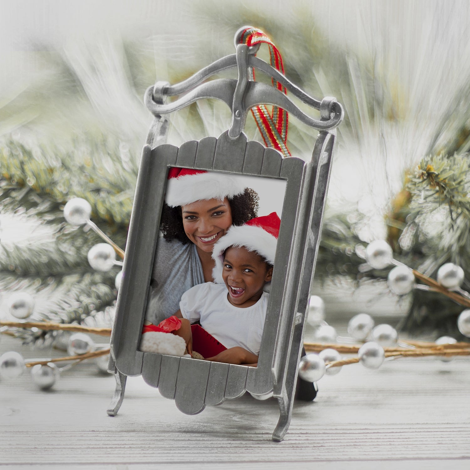 A pewter sled-shaped photo ornament and picture frame adorned with a red, green, and gold ribbon displayed on a table on a festive lifestyle background.