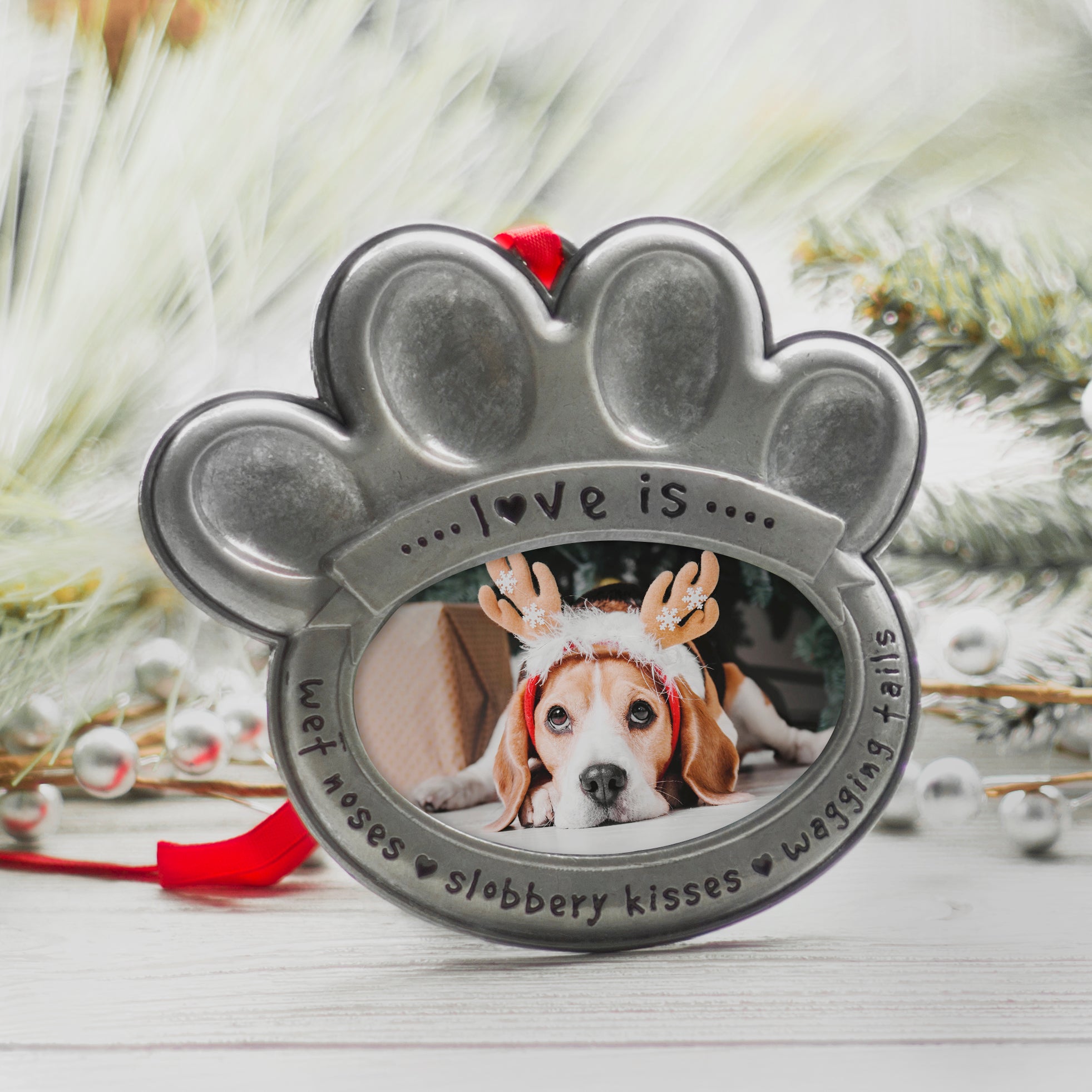 A pewter photo ornament in the shape of a paw print with the text 'love is wet noses, slobbery kisses, wagging tails" displayed on a table of a festive lifestyle background.