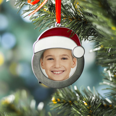 A round pewter photo ornament with a Santa hat, a black velvet easel, and a red velvet ribbon hanging from a Christmas tree on a festive lifestyle background.