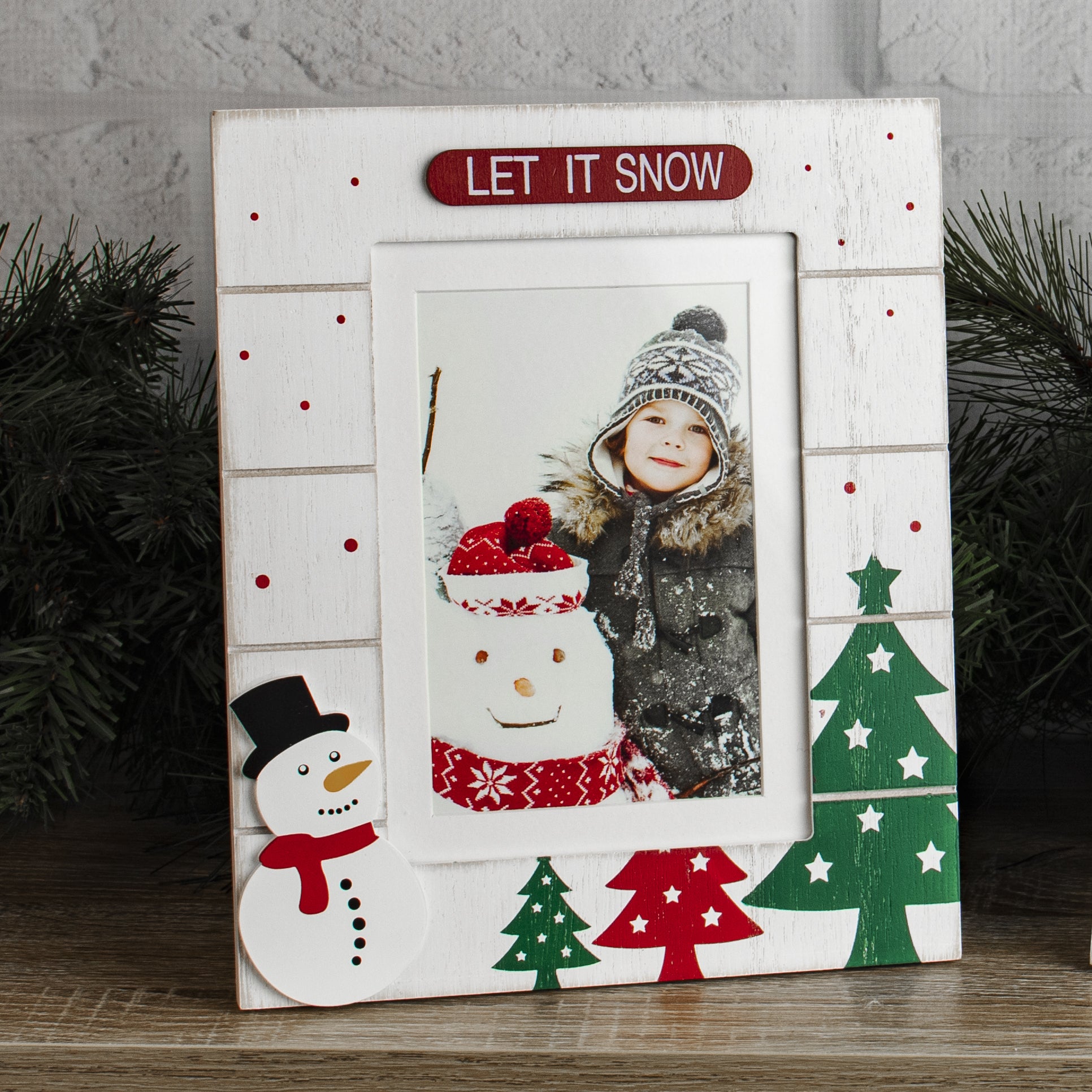 A white, wooden picture frame with a 'LET IT SNOW' sign at the top, and designs featuring Christmas trees, and and a snowman on a festive lifestyle background.