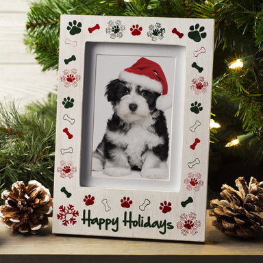 A white wooden picture frame with the phrase "Happy Holidays" at the bottom with a border of paw prints, bones, and snowflakes displayed on a festive lifestyle background.