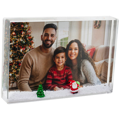 A clear acrylic snow globe frame with a white faux-snow base, displaying a professional Christmas portrait. A small green Christmas tree and a Santa Claus figure, rest in the snow. Used for Santa photo packages by professional photographers and sold in bulk at wholesale prices.
