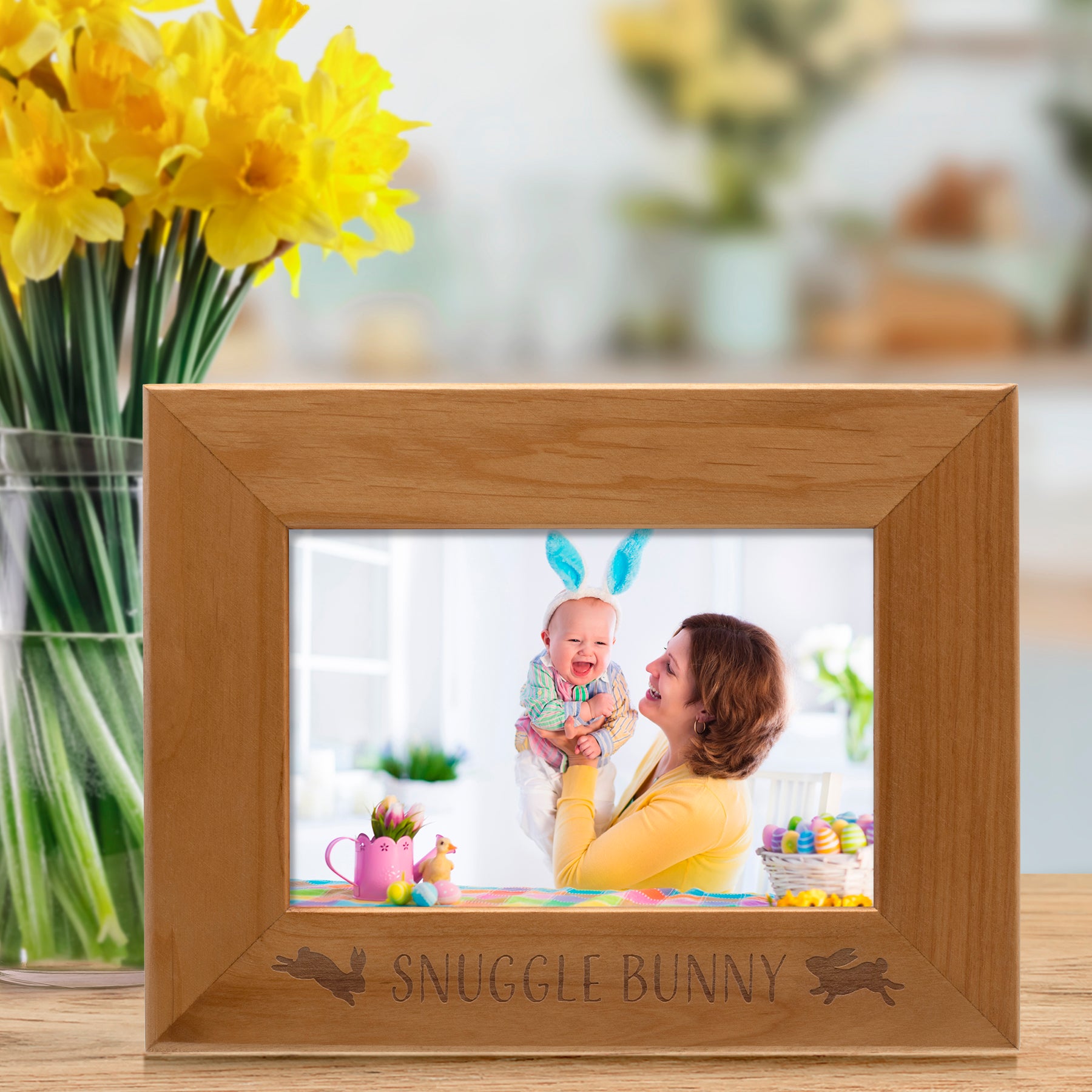 Wooden photo frame with 'Snuggle Bunny' engraving displaying a photo of a woman and child, next to a vase of yellow flowers.