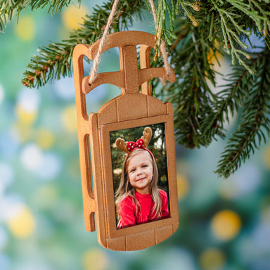 A wooden sled-shaped photo ornament seen hanging on a Christmas tree by a loop of twine.