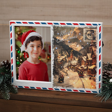 A plastic picture frame designed to resemble a postcard, with a vintage Christmas-themed image on the right and a space for a photo on the left. The frame features mini fiber optic lights to mimic snow and stars. This frame is shown sitting on a shelf or mantel. 