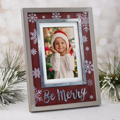 A Christmas-themed wooden picture frame with a gray border and reddish/brown insert. It features white snowflakes in varying sizes in the background and the phrase 'Be Merry' in silver at the bottom shown with a festive back drop.